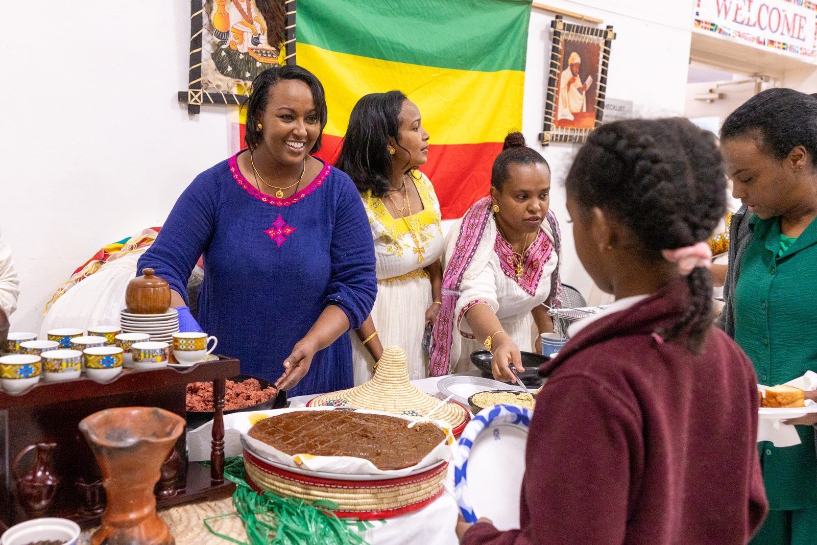 People at a food stall with jars, bread, and bowls, standing before a green, yellow, and red flag.