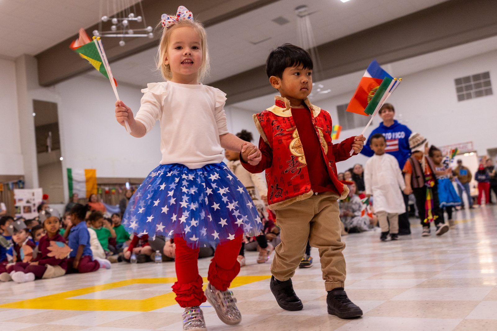 Two children in festive clothes walk hand in hand at a school cultural parade, each holding a small flag.