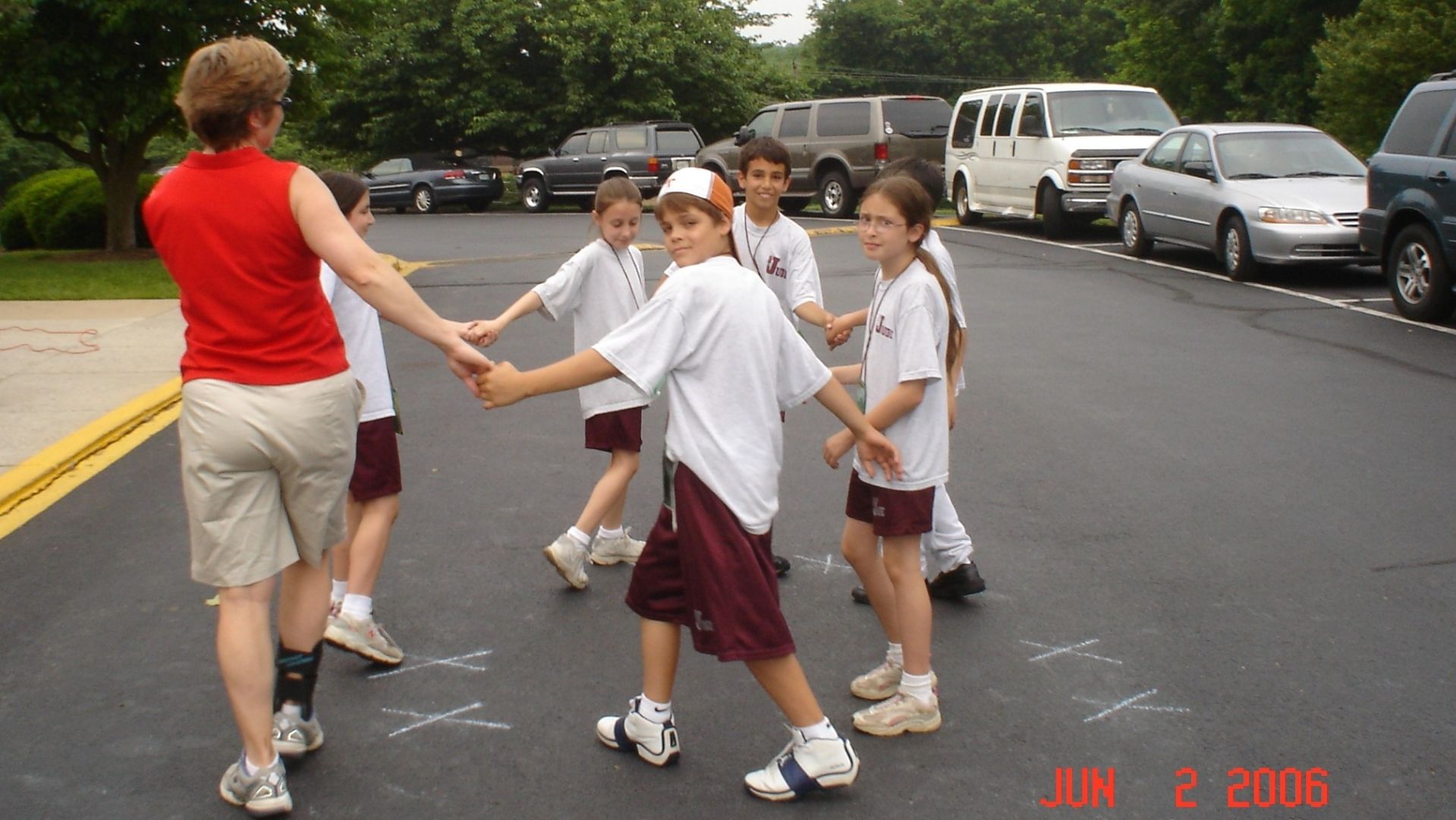 Children holding hands and walking across a street near parked cars, with an adult leading them