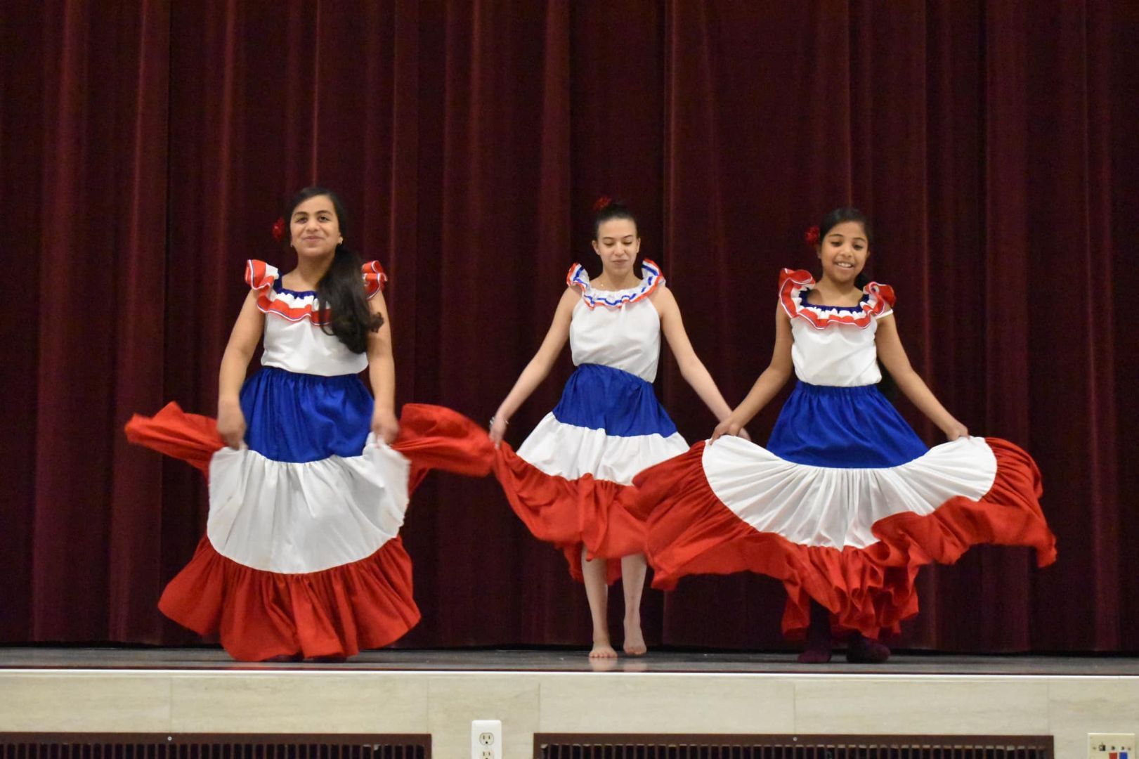Three dancers in red, white, and blue dresses pose onstage in front of a burgundy curtain.