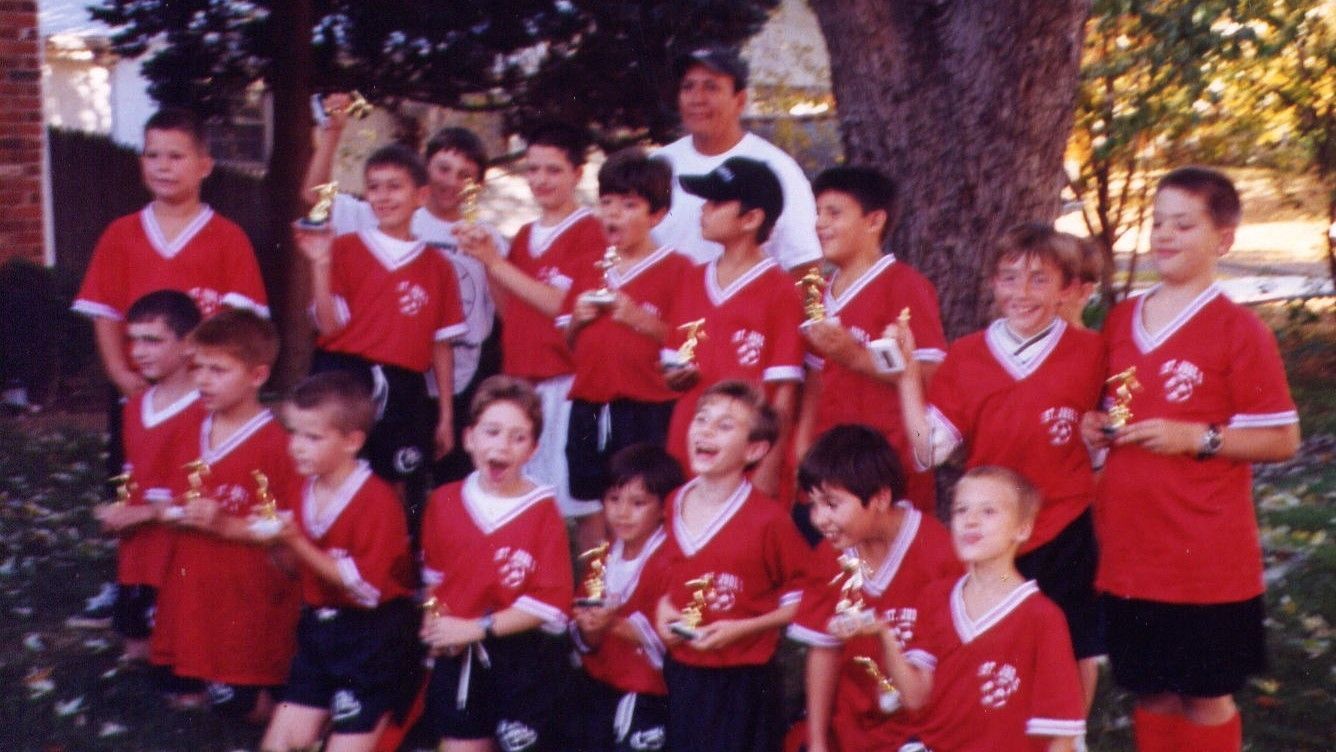 Group of children in red sports jerseys posing outdoors, some holding certificates and trophies.