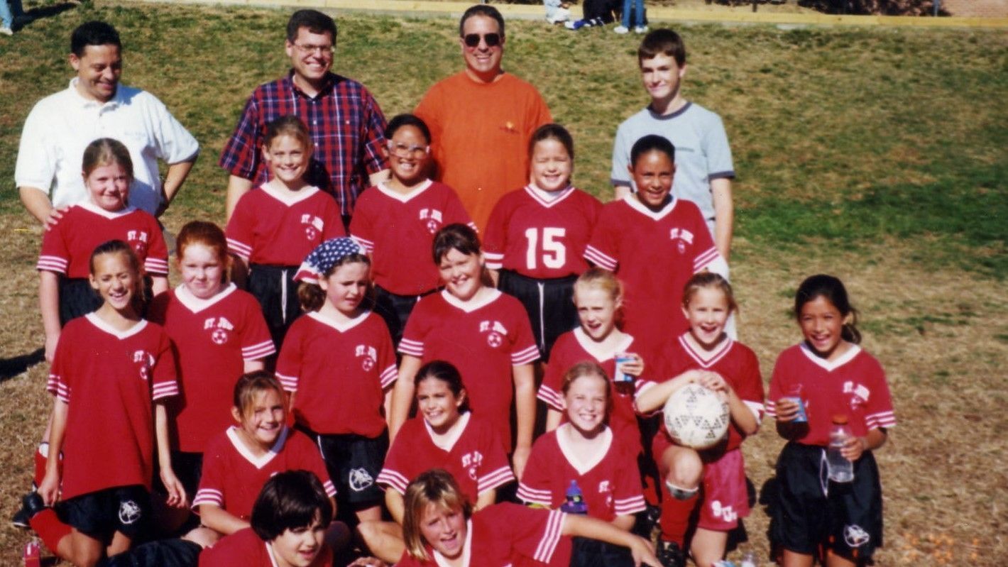 Youth soccer team posing outdoors, wearing red uniforms with coaches behind them.
