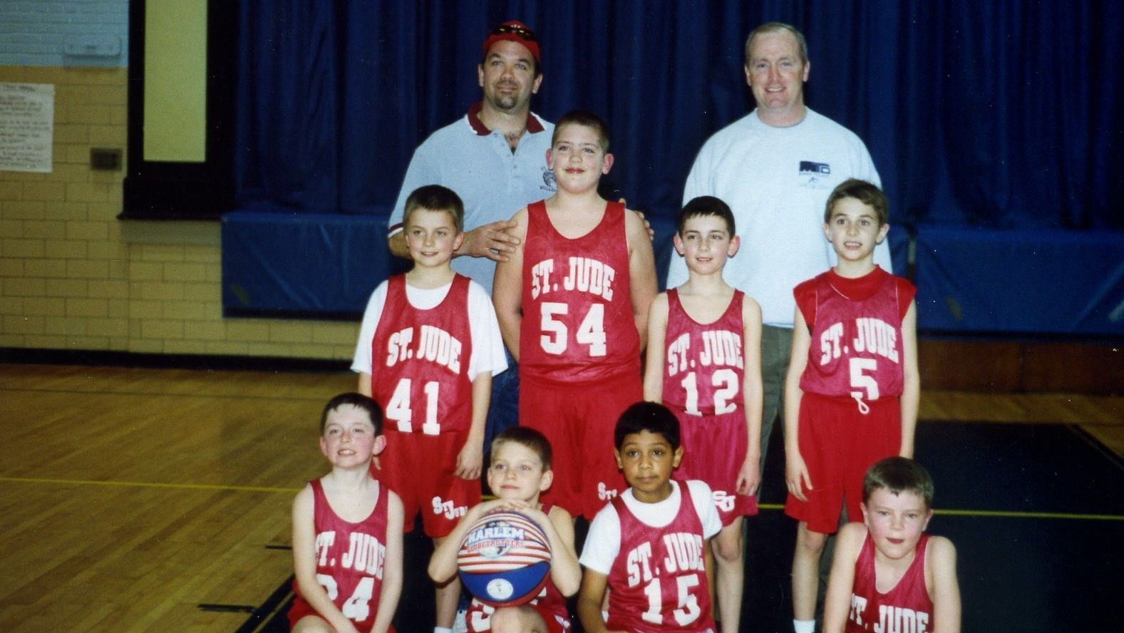 Youth basketball team posing in red jerseys with coaches in a gym.