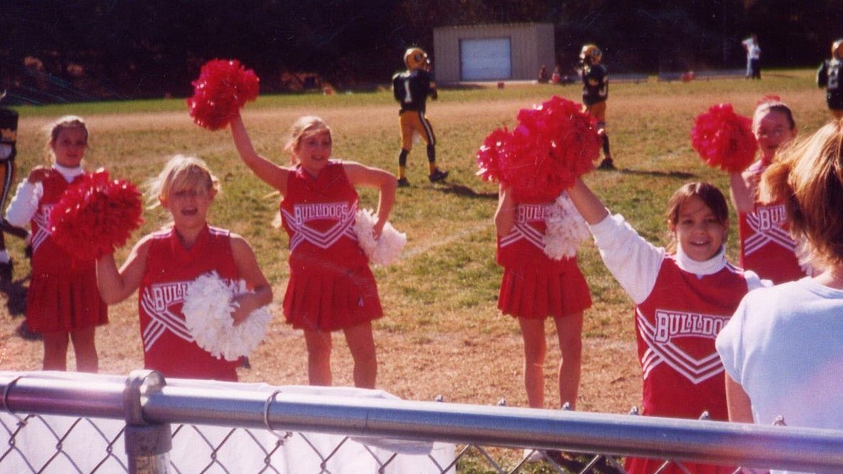 Cheerleaders in red uniforms holding pom-poms on a sports field behind a chain-link fence