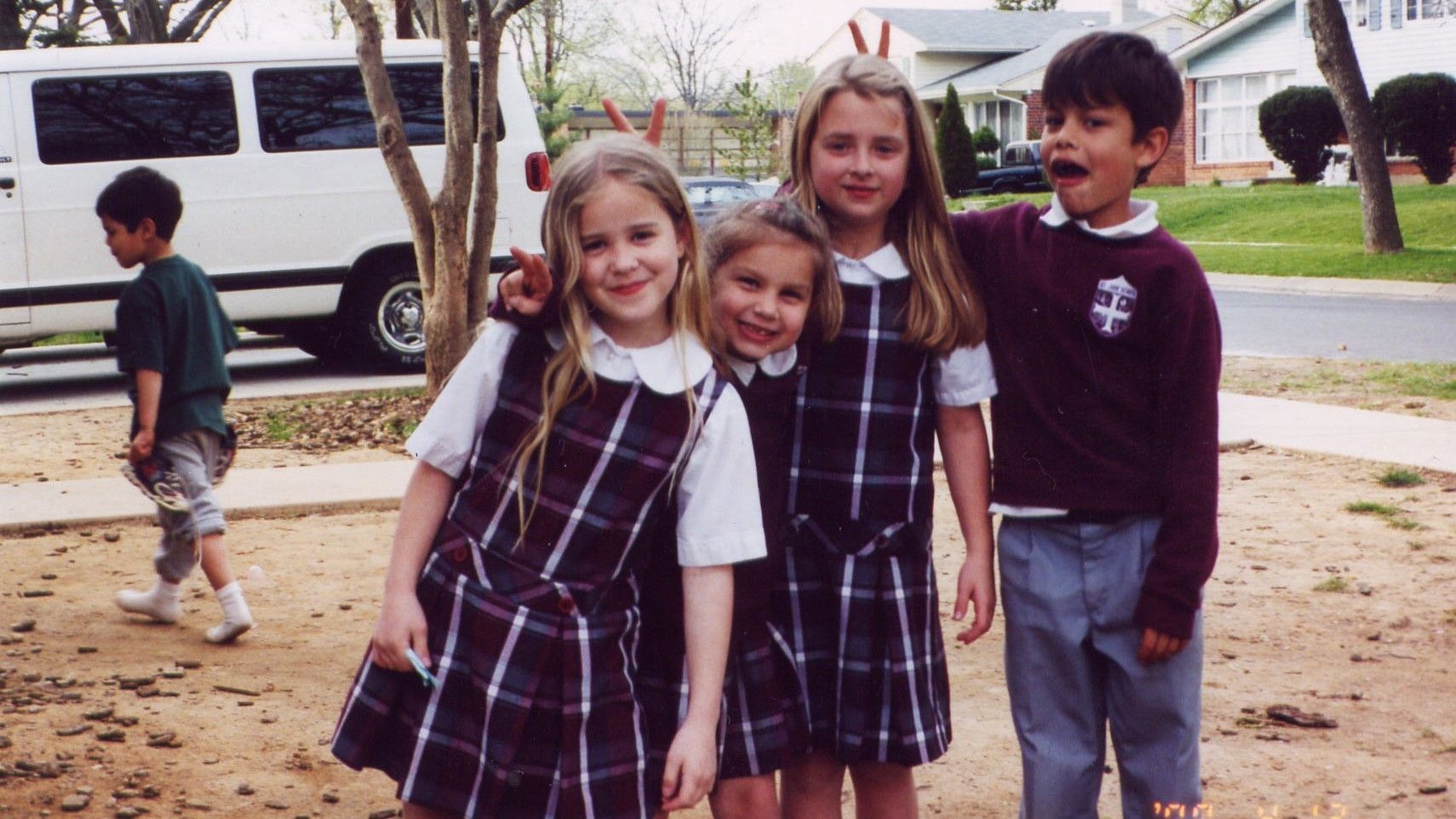 Four smiling children stand outdoors near a white van, with another child walking in the background.