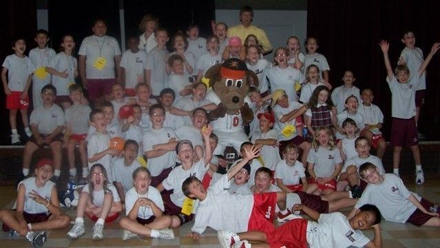 Large group posing indoors with a costumed mascot in the center, many wearing white shirts and yellow sun shapes.