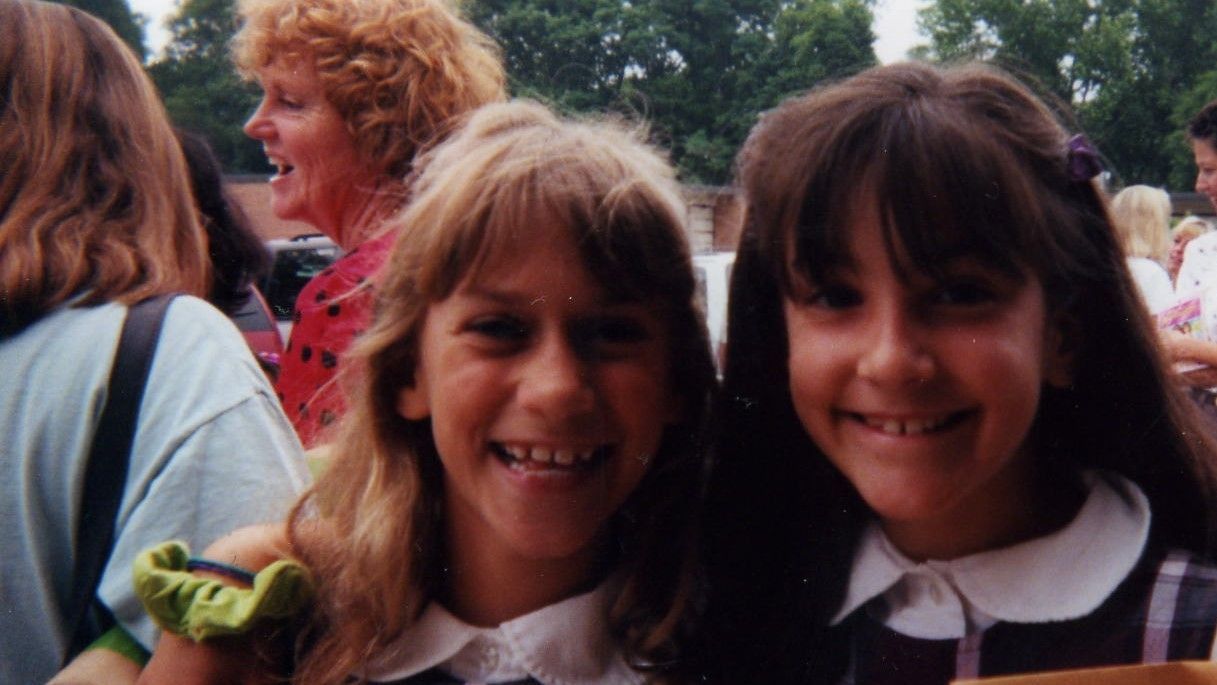 Two smiling children posing outdoors in front of a crowd, with one wearing a dark sweater and white collar.