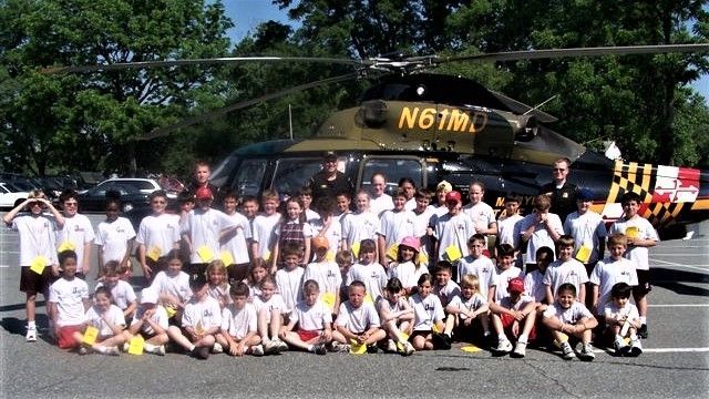 Large group posing in white shirts in front of a black helicopter on a sunny roadside