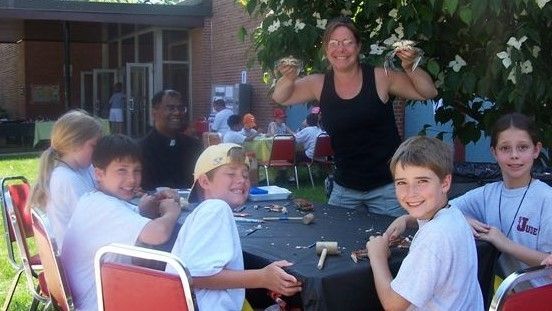 People seated outdoors at a table while a woman in a black top stands behind them, smiling.