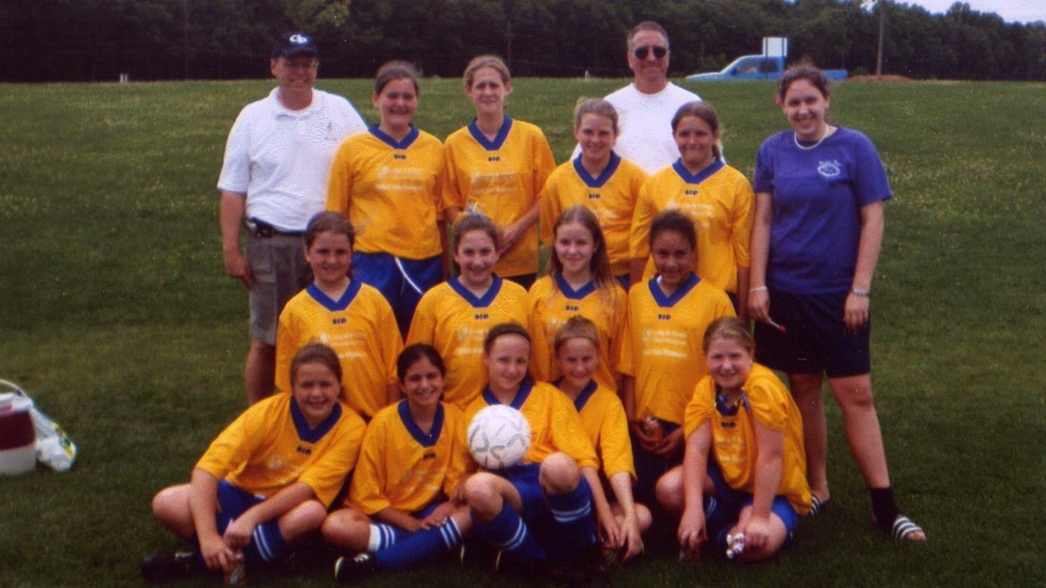 Youth soccer team posing on a grassy field, wearing yellow and blue uniforms, with coaches standing behind.