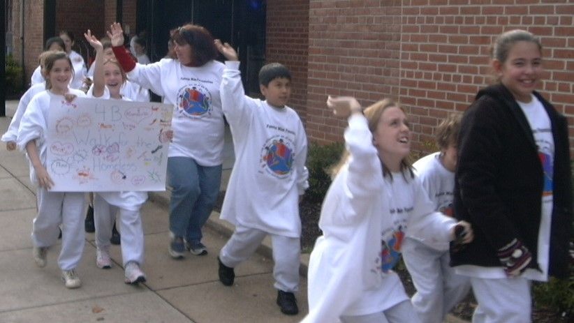 Children in white outfits march with signs in a school parade outside a brick building
