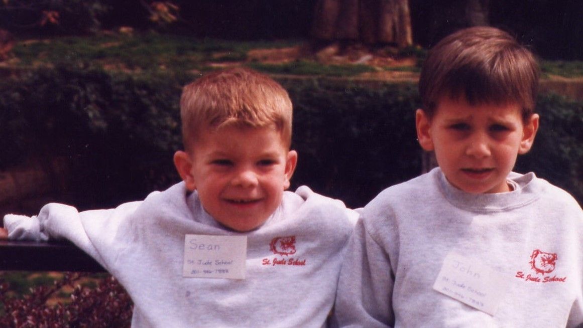 Two children in white sweatshirts smiling outdoors, with arms around each other.