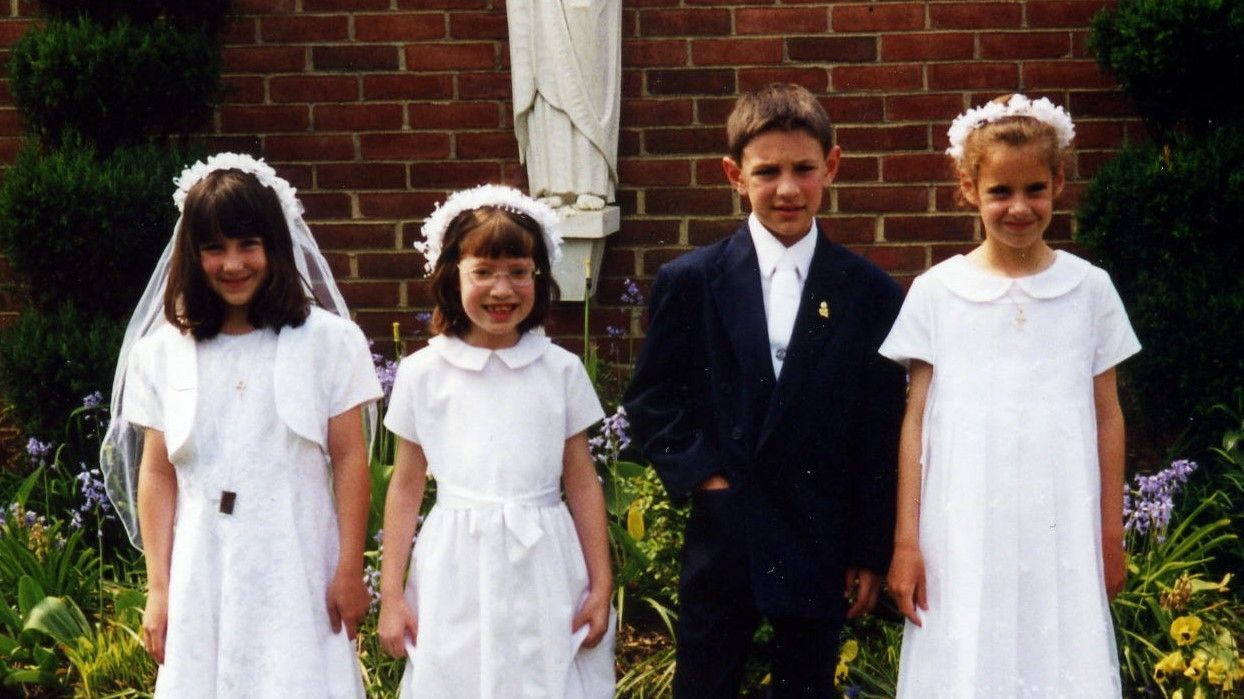Four children dressed for a formal event standing outdoors in front of a brick wall and flowers.