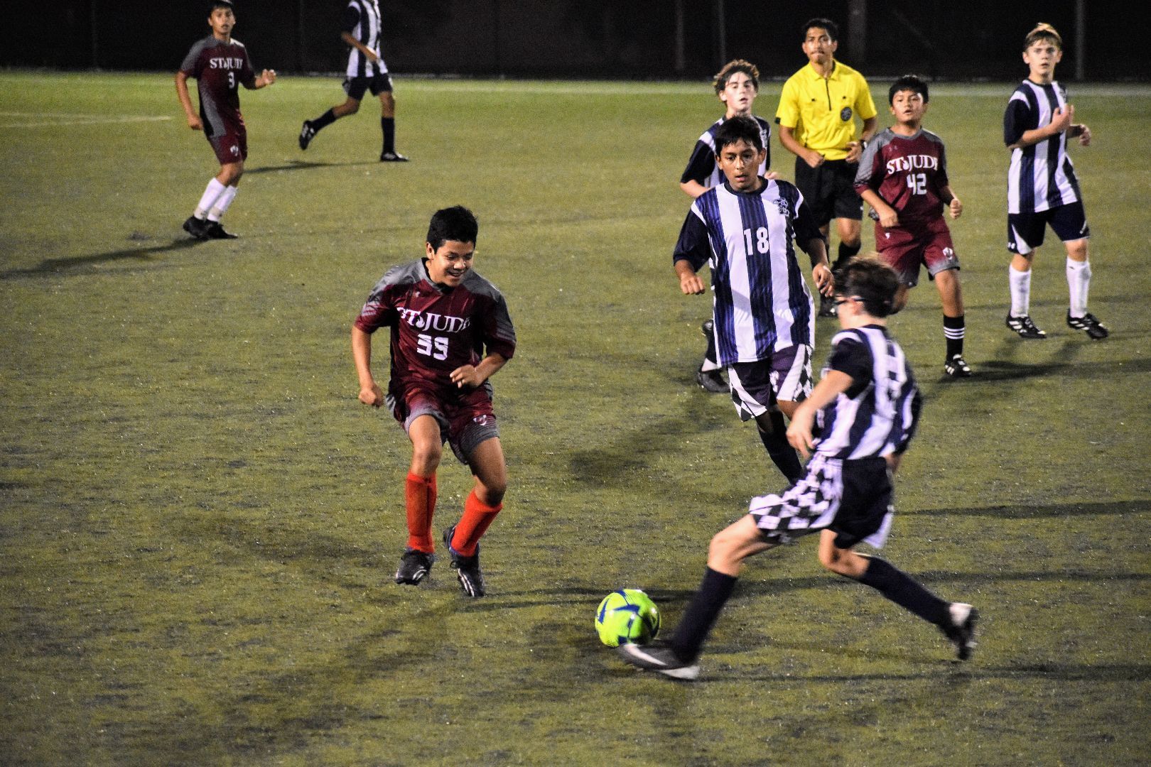 Soccer players in black-and-white stripes and dark uniforms chase the ball on a lit field at night