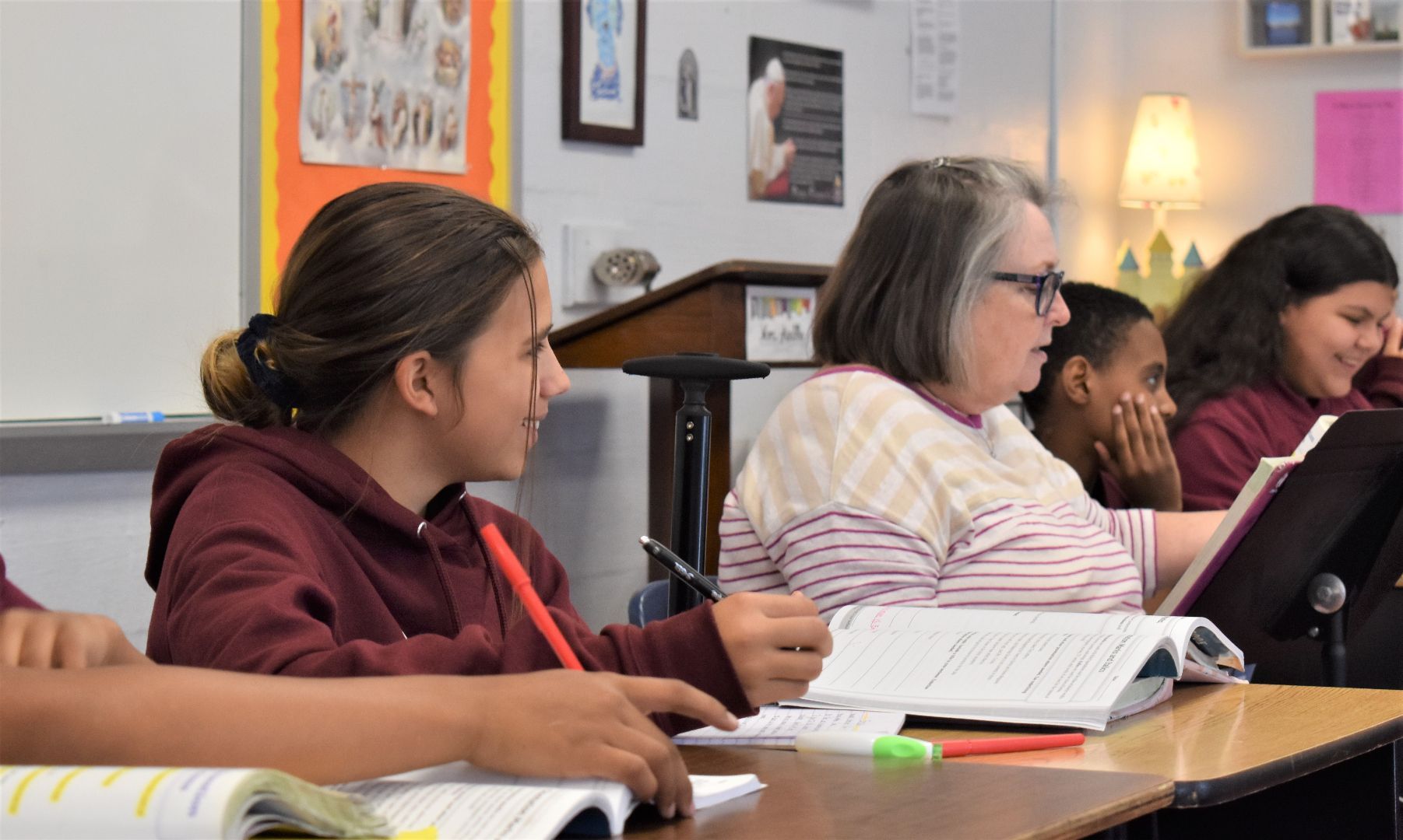 Students and a teacher seated in a classroom, listening and reading from open books at a desk.