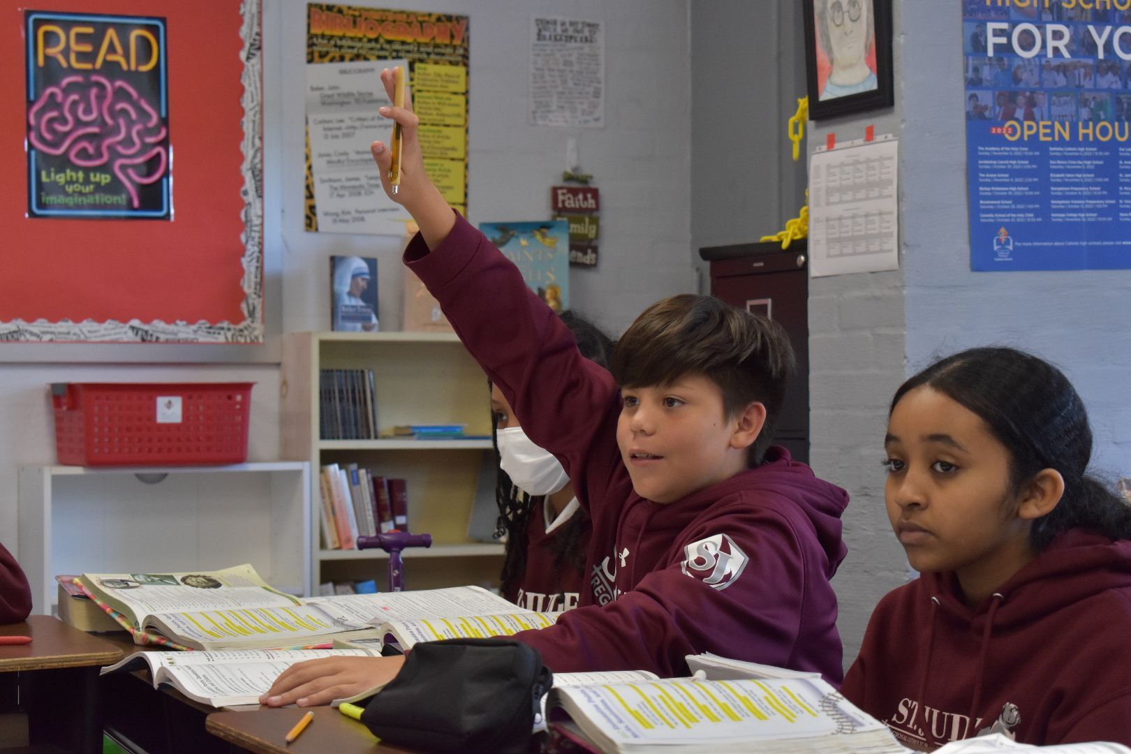 Two students in maroon uniforms at desks, one raising a hand in a classroom with posters and bookshelves.