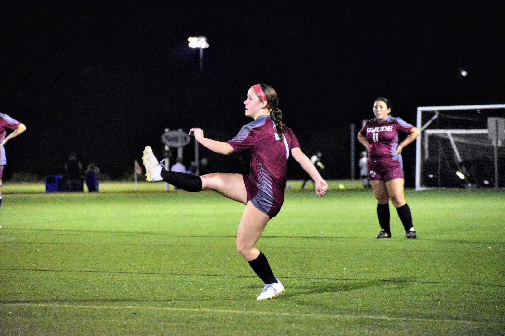 Soccer player in maroon jersey kicks ball on lit field at night, with goal and teammates in background