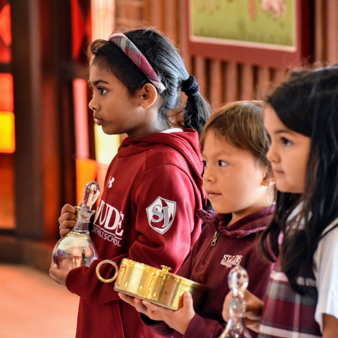 Children in maroon school uniforms holding small golden cups indoors