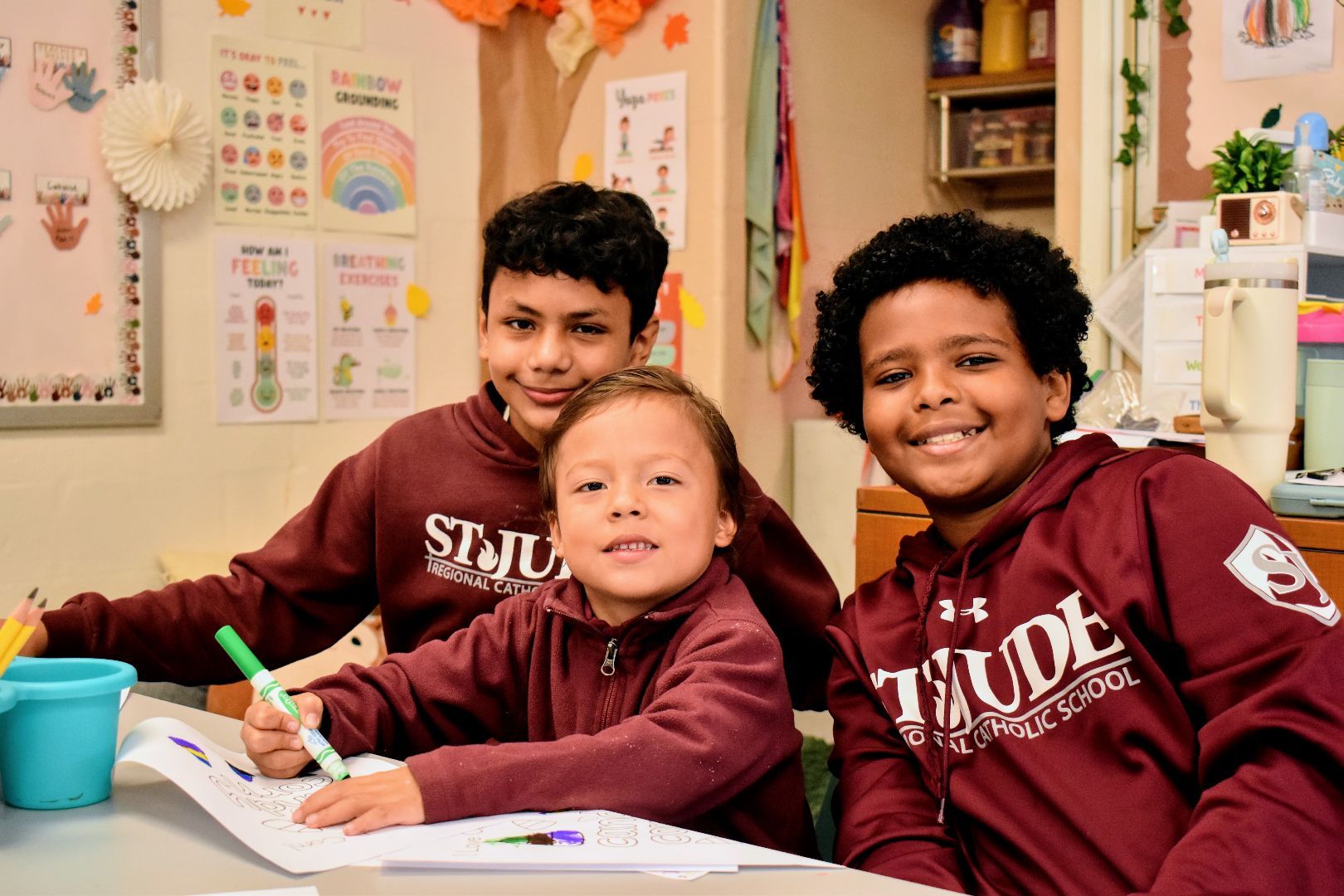 Three children in maroon sweatshirts sit at a table, smiling and drawing in a decorated classroom.