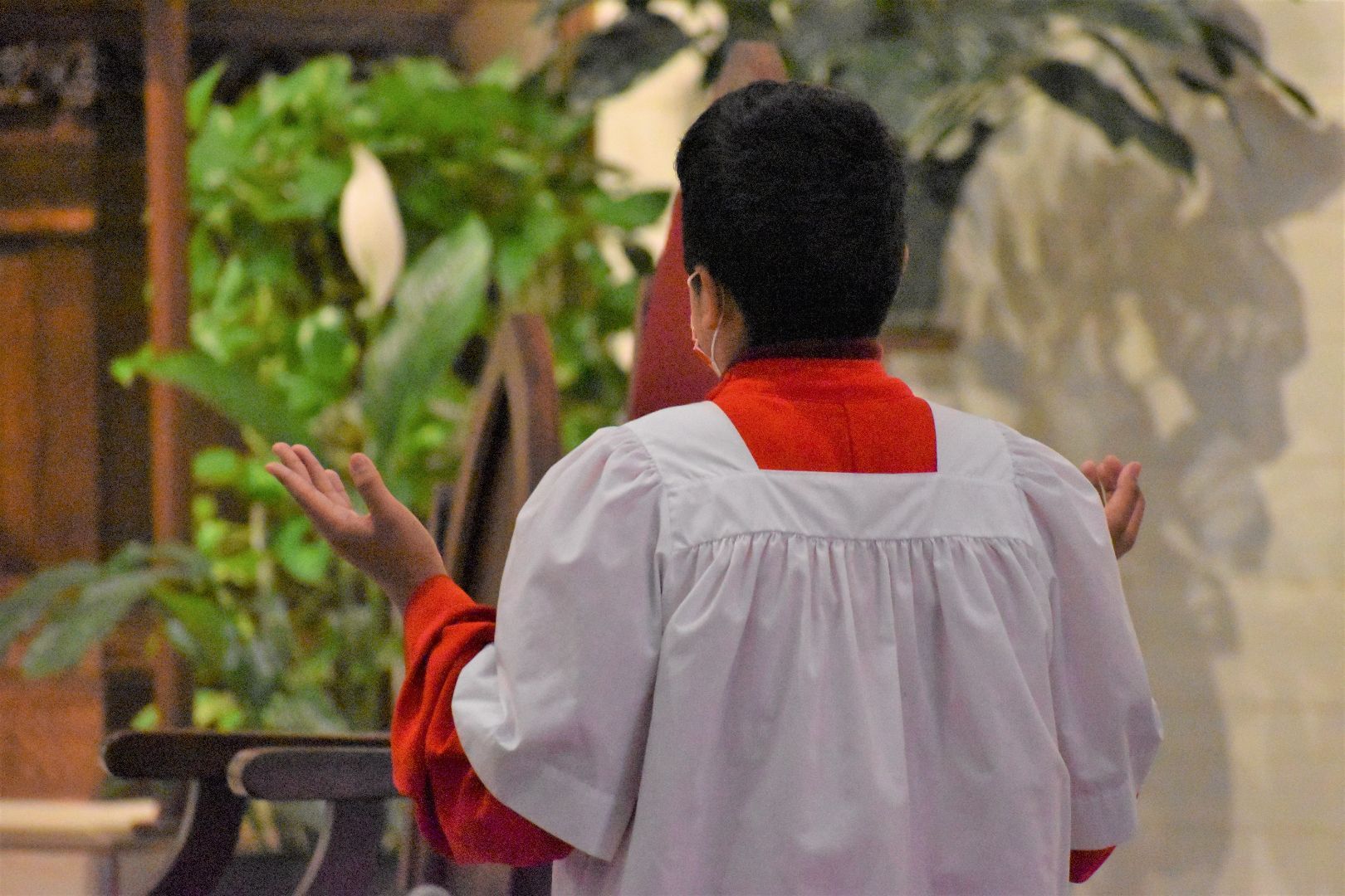 Person in a white and red robe stands with raised hands in a church, seen from behind.
