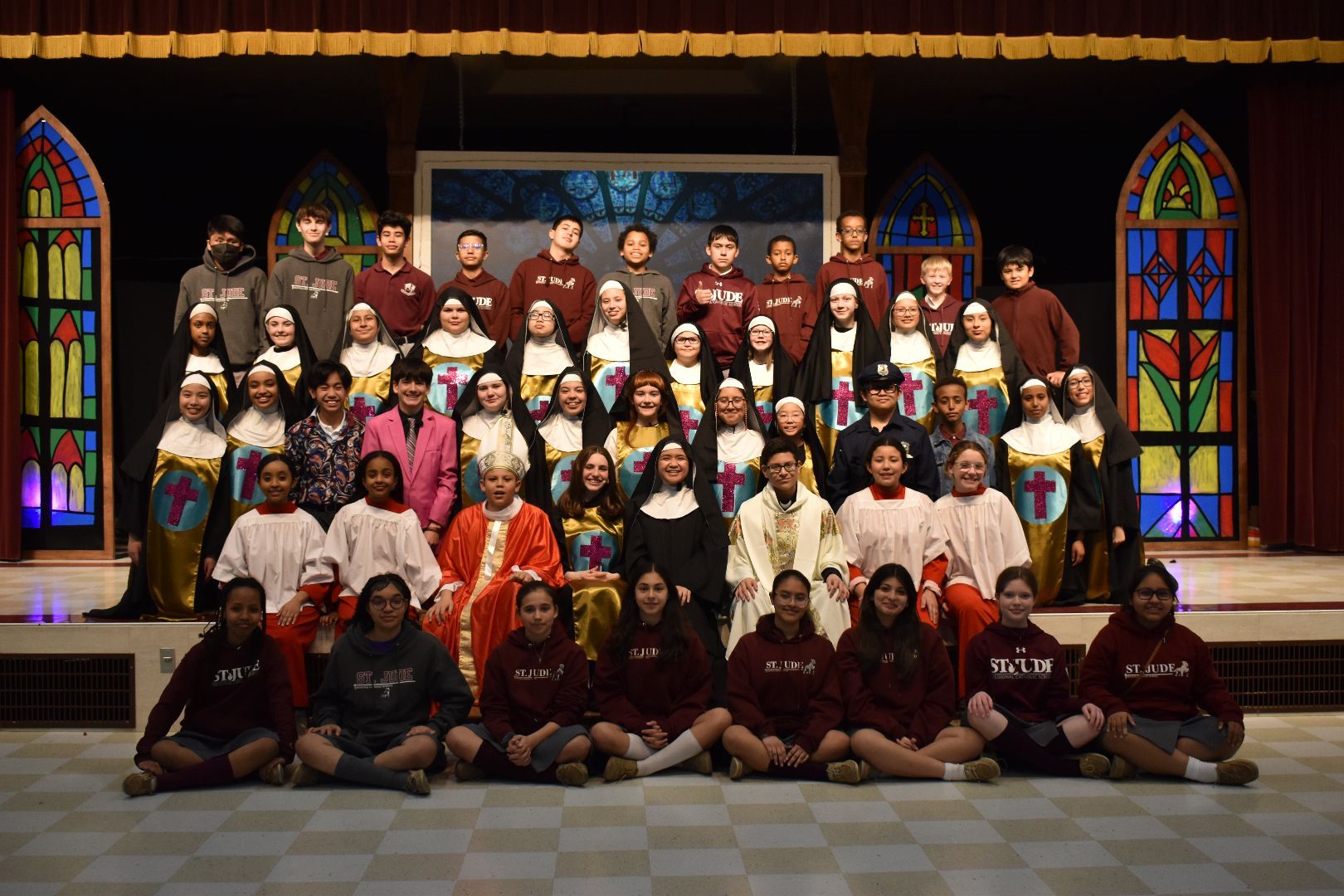 Large group posing on a stage in colorful costumes with stained-glass-style backdrops