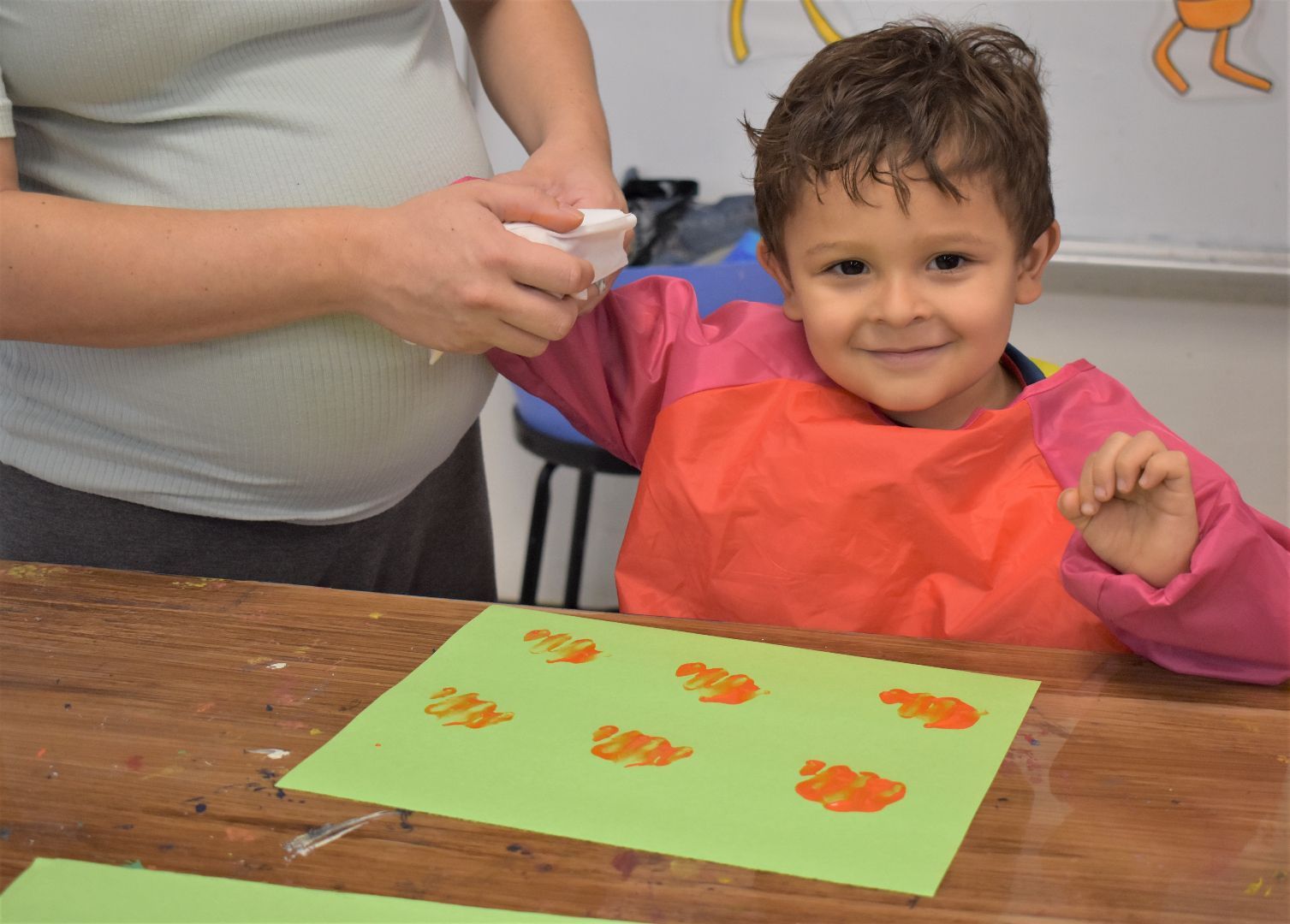 Child in smock smiling at craft table while an adult applies paint to their arm
