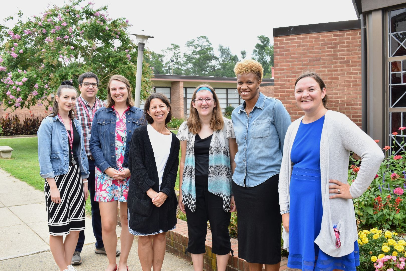 Seven teachers stand smiling outdoors in front of a brick building and flowers.