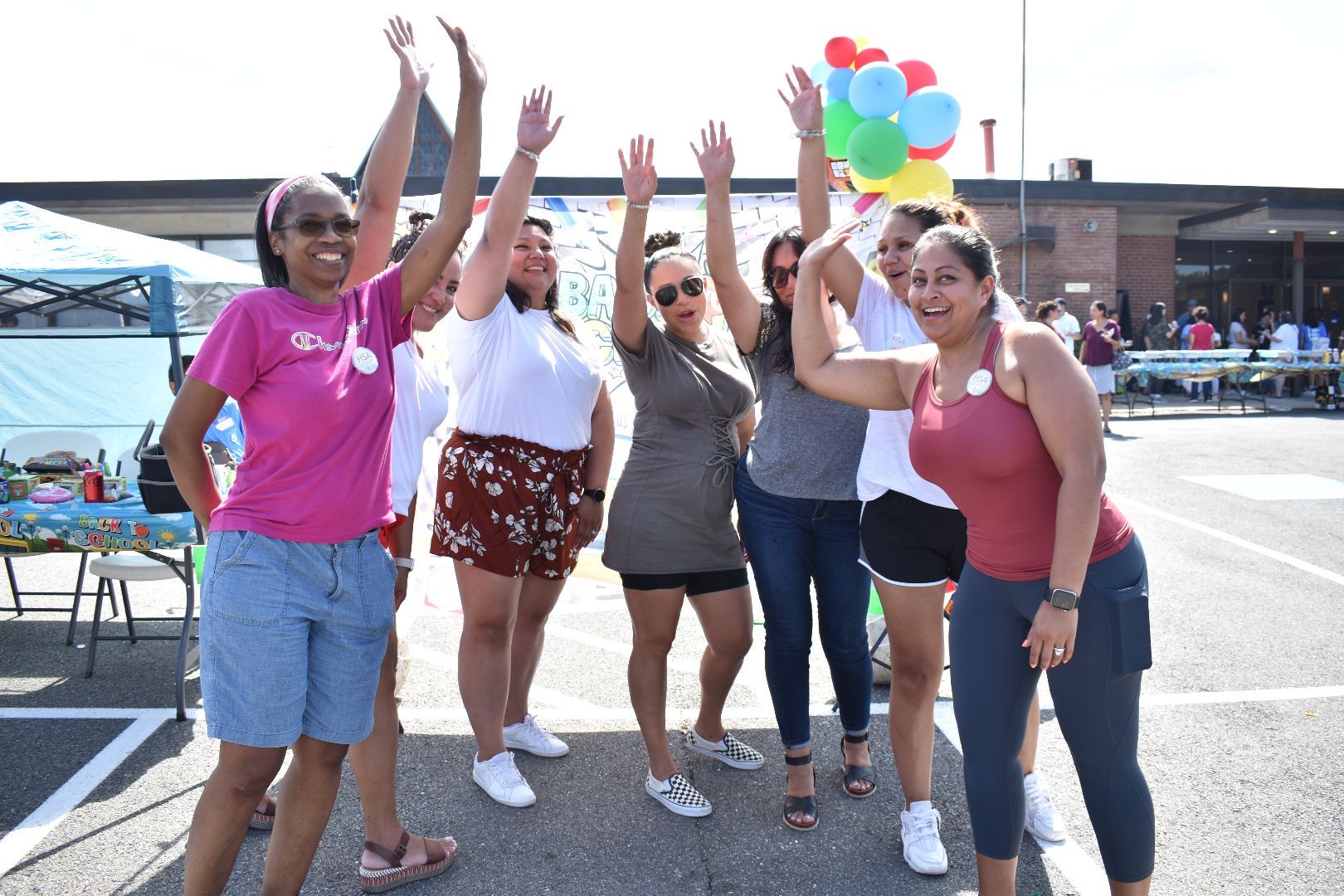 Six people raising their arms outdoors, smiling in a parking lot with colorful balloons in the background