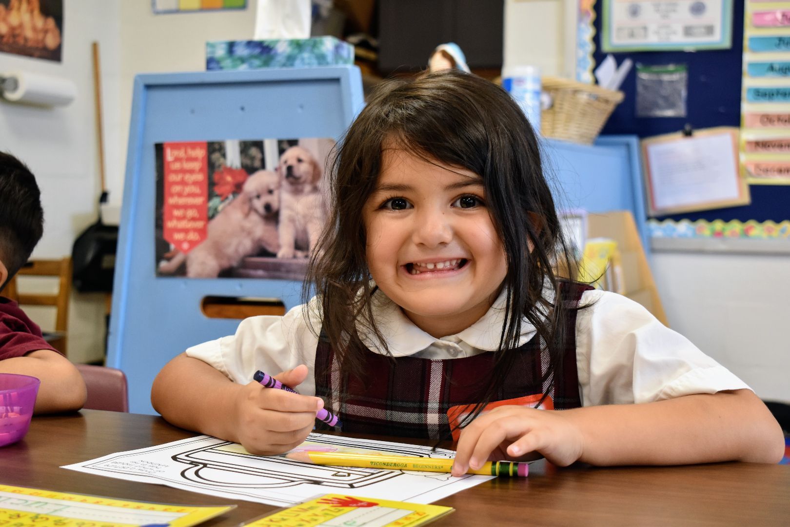 Smiling child coloring at a classroom desk with crayons and a worksheet