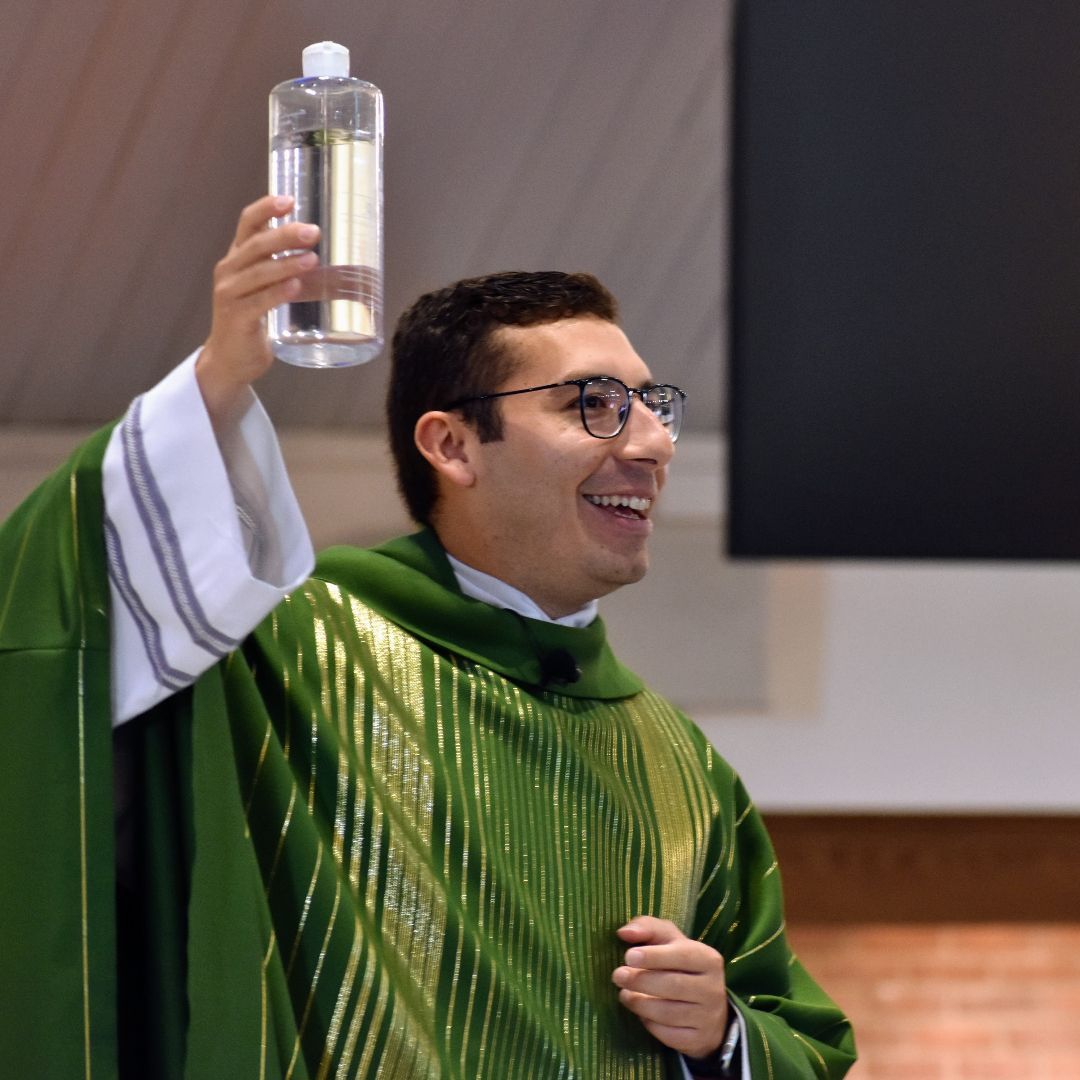 Priest in a green vestment raises a clear bottle during a church service