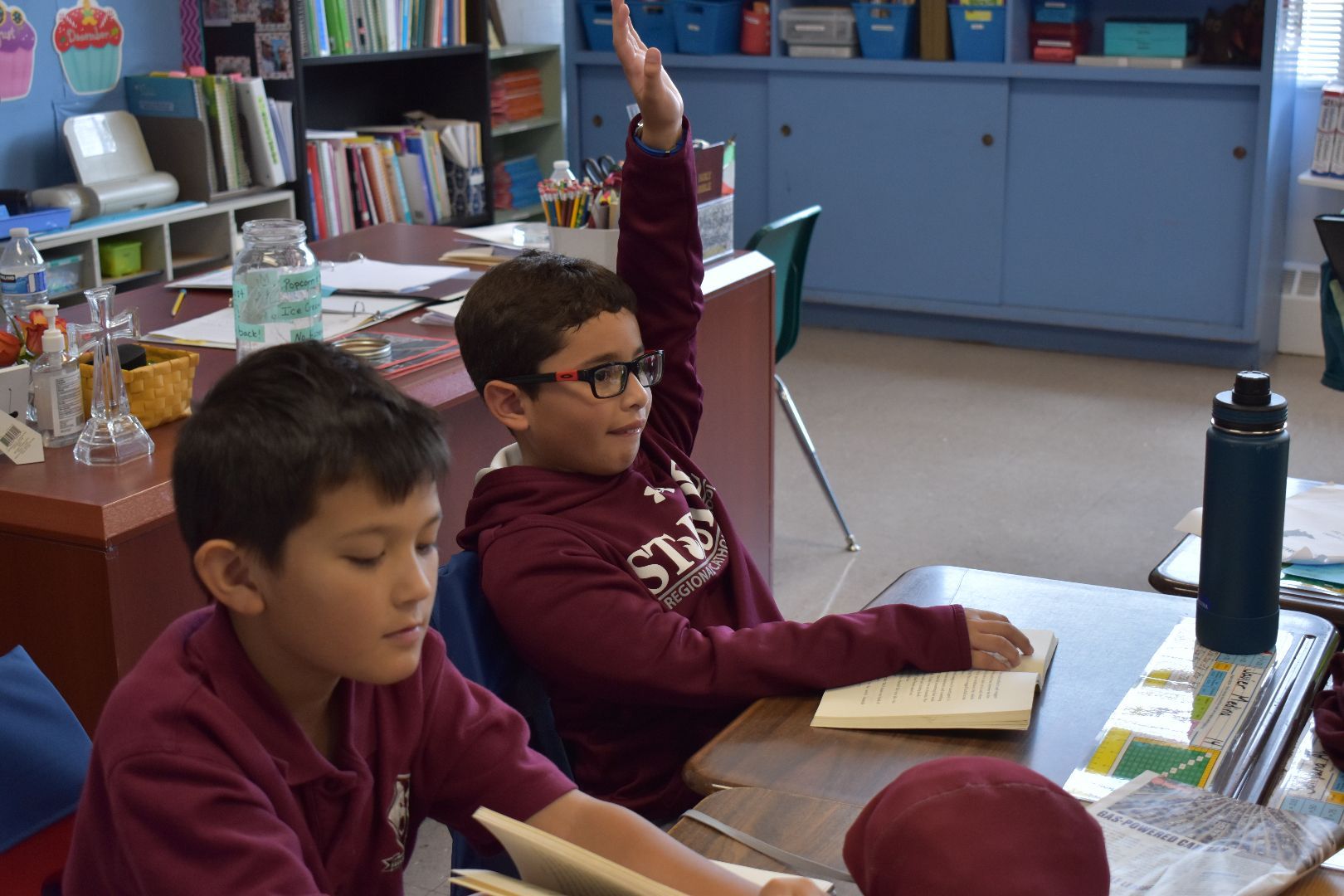 Students seated at desks in a classroom, one raising a hand while another looks down at a notebook.