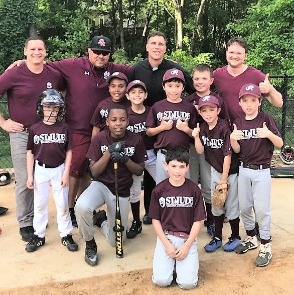 Youth baseball team and coaches posing outdoors in maroon jerseys, with bats and gloves, in front of trees.