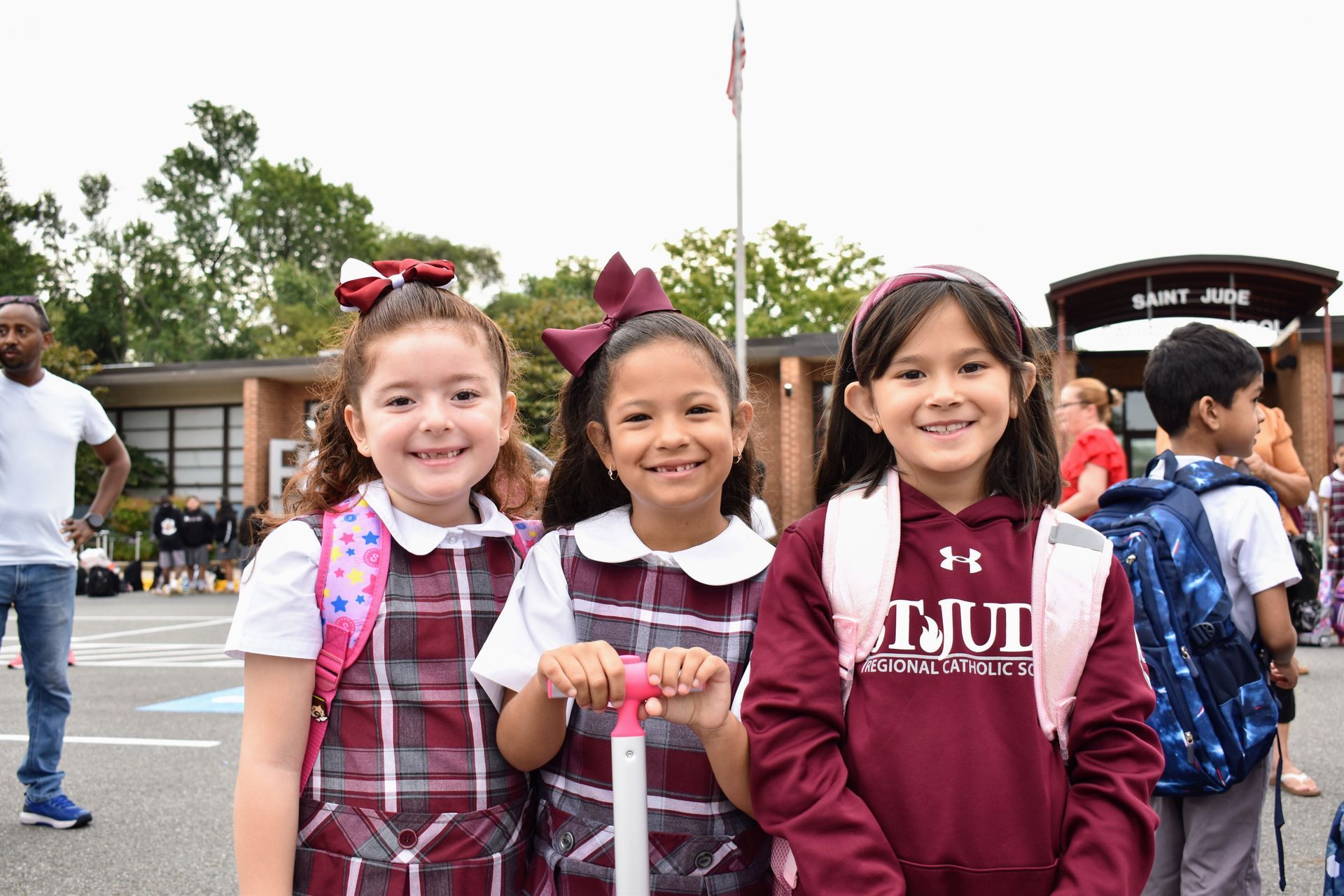 Three smiling children holding lanyards stand outdoors near a school building.