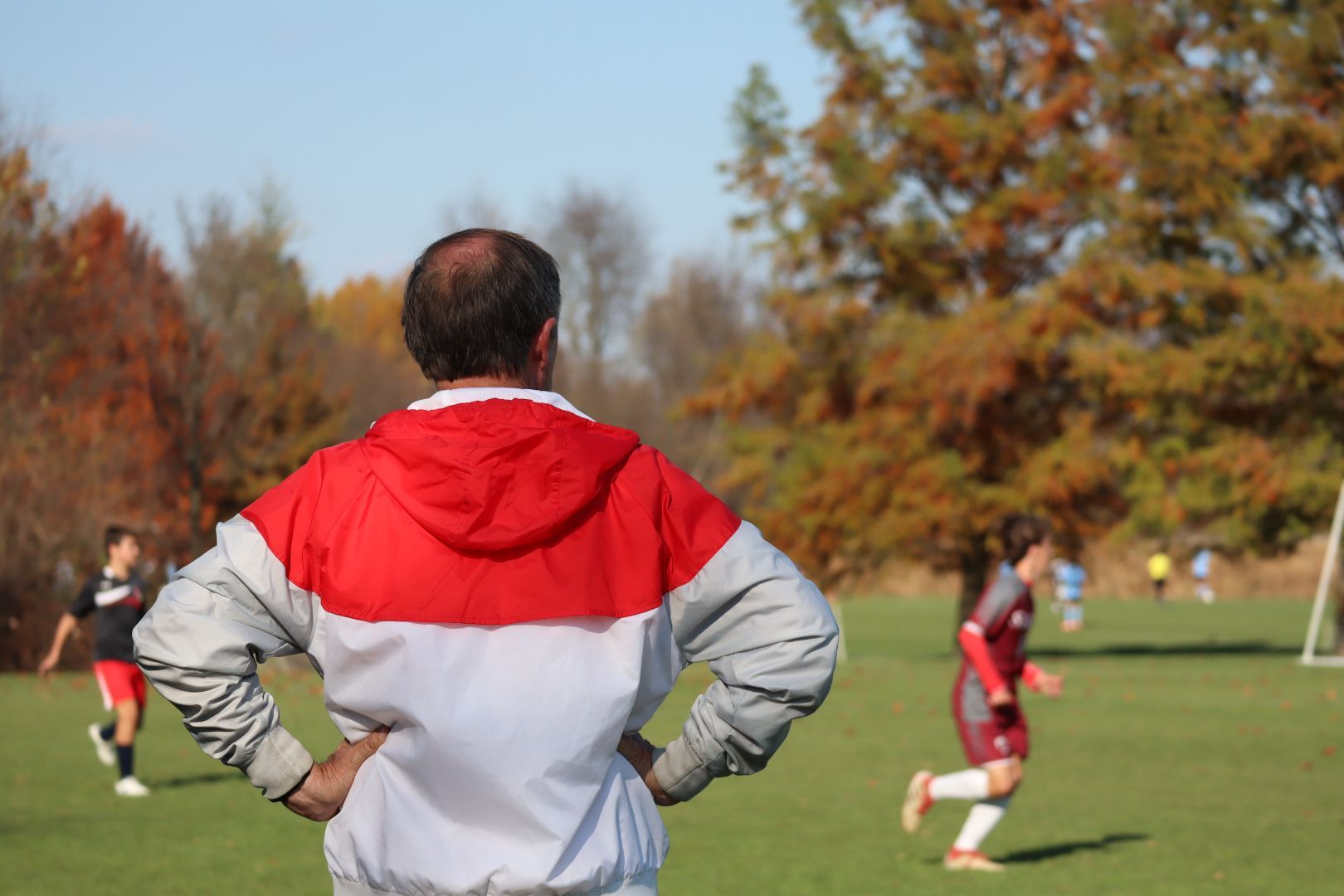 Coach in red and white jacket watching a soccer game on a grassy field with players in the background