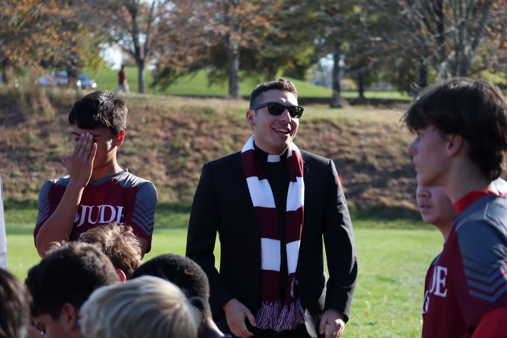 Coach in sunglasses and scarf talks to players on a sunny sports field.