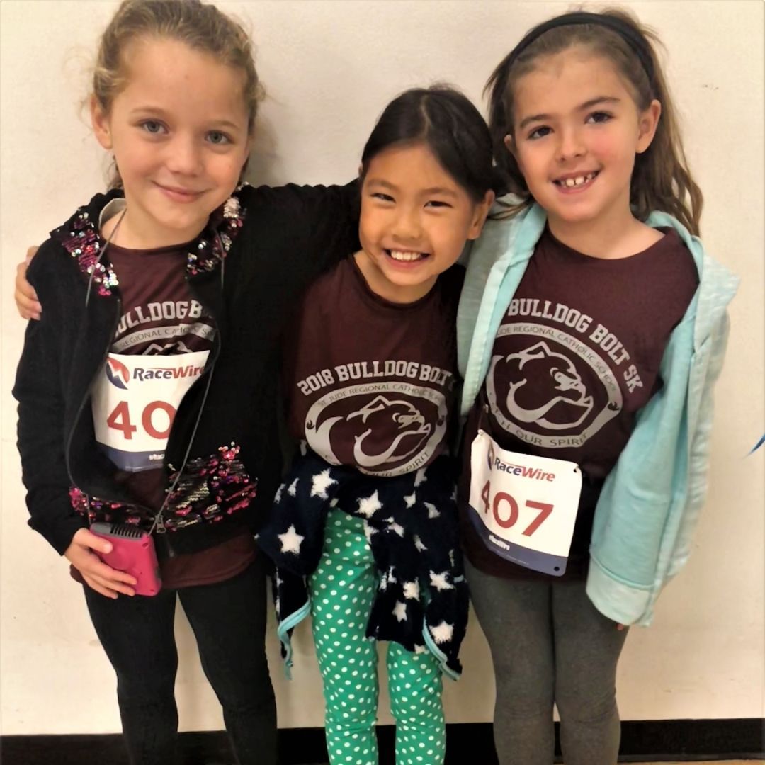 Three girls in matching shirts and race bibs stand arm-in-arm indoors, smiling at the camera.