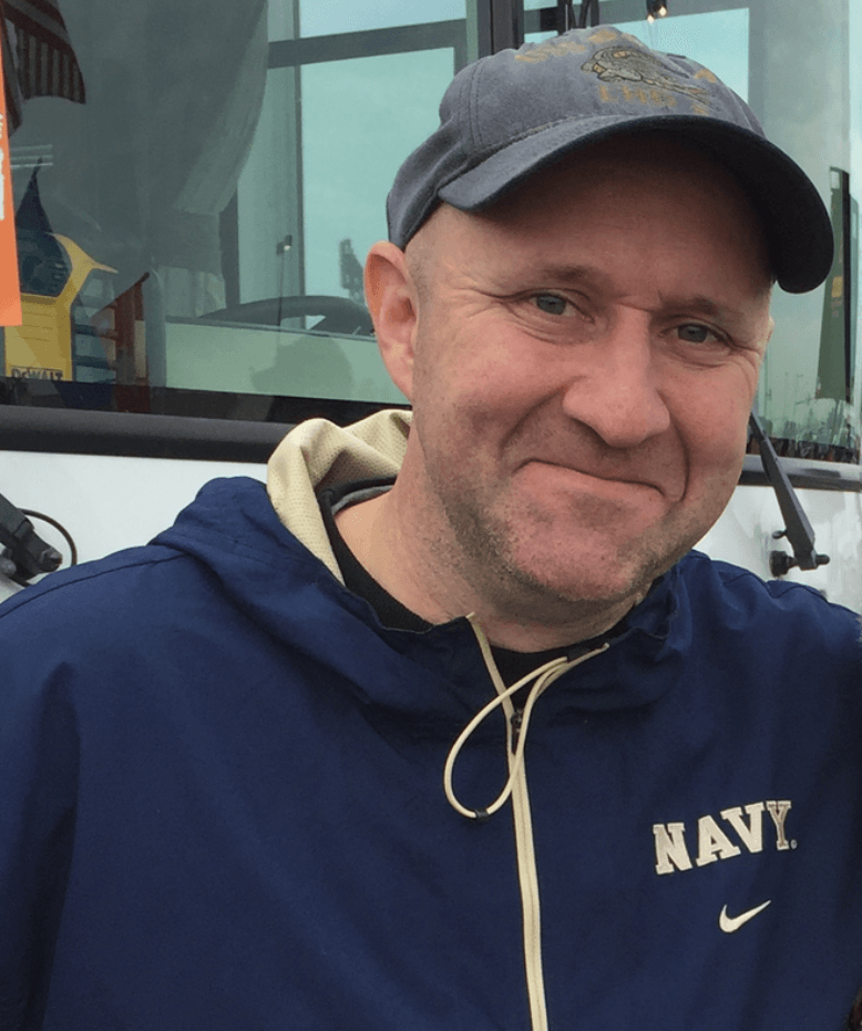 Smiling person in a navy hoodie and cap standing in front of a bus.