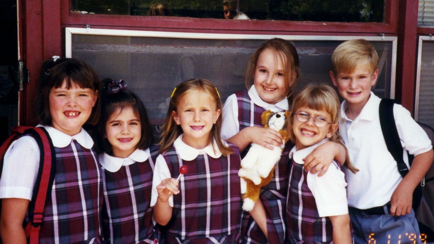 Six smiling children in plaid school uniforms standing together outdoors