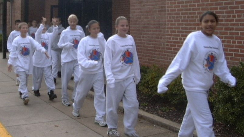 Children in white sweatshirts and pants walking in a line along a sidewalk beside a brick building