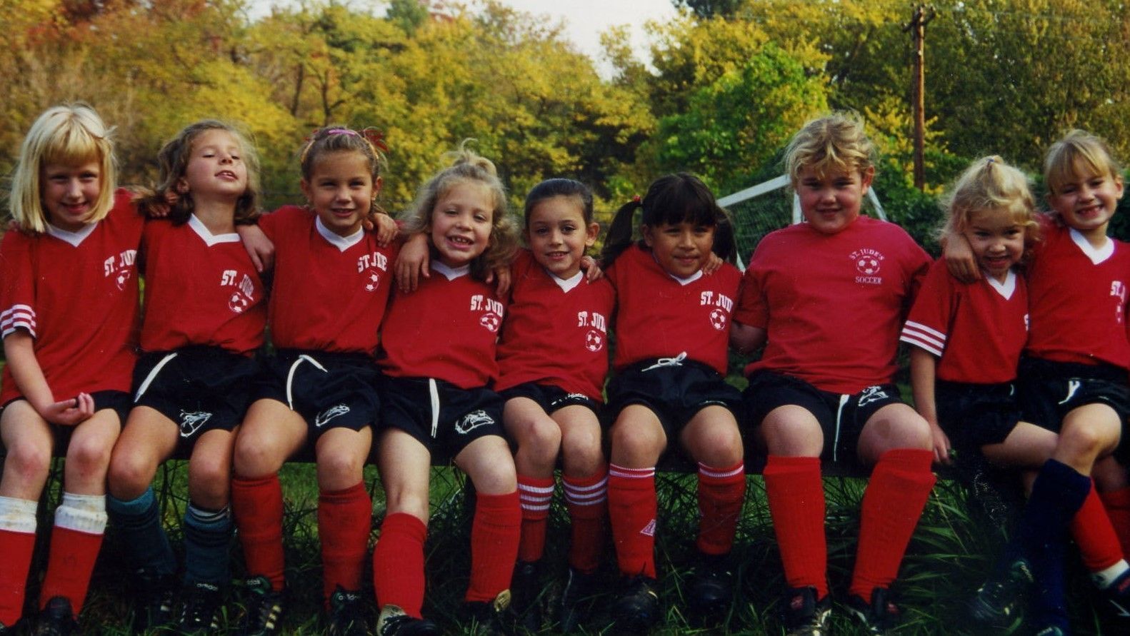 Children in red sports uniforms sitting on a bench outdoors, smiling in a team lineup