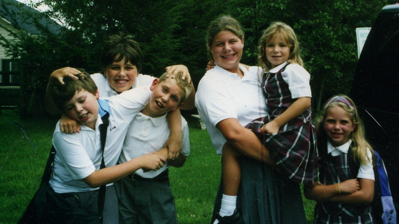 Five children in white shirts pose and smile outdoors on a grassy lawn.
