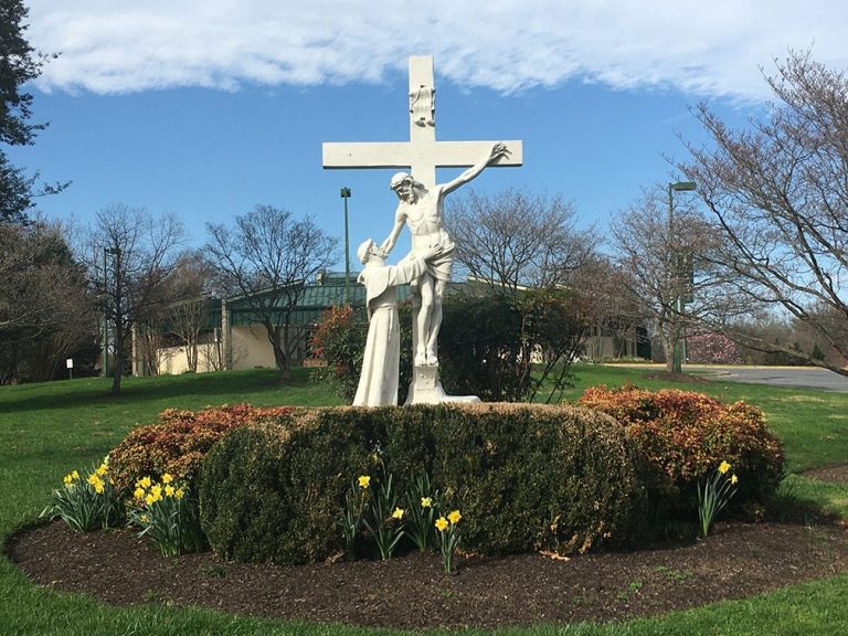 White religious statue with cross in a flower bed, set in a grassy park under a blue sky