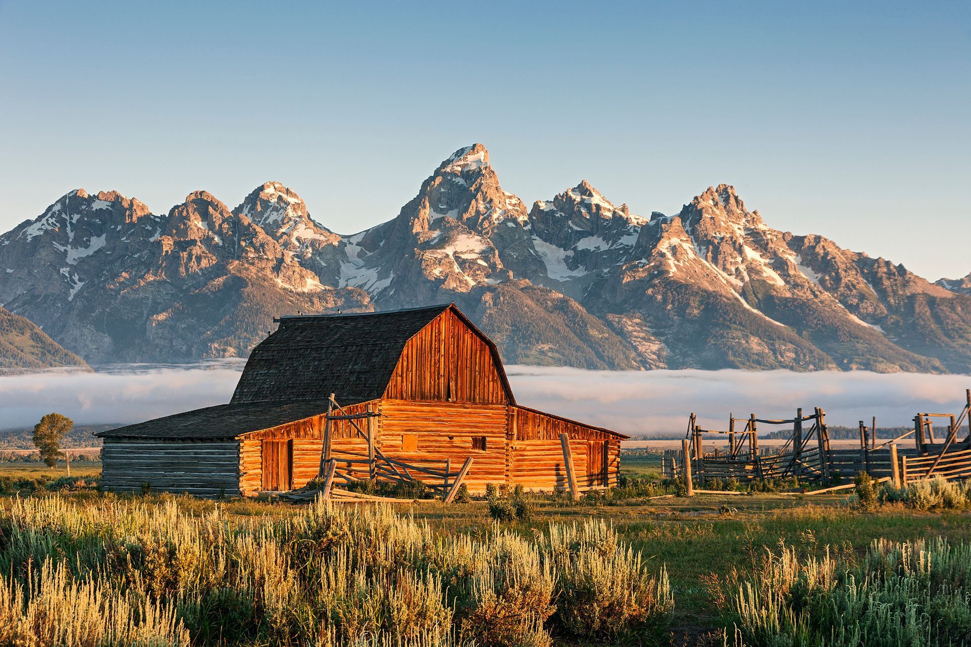 Rustic barn in a grassy field with snow-capped mountains under a clear sky at sunset