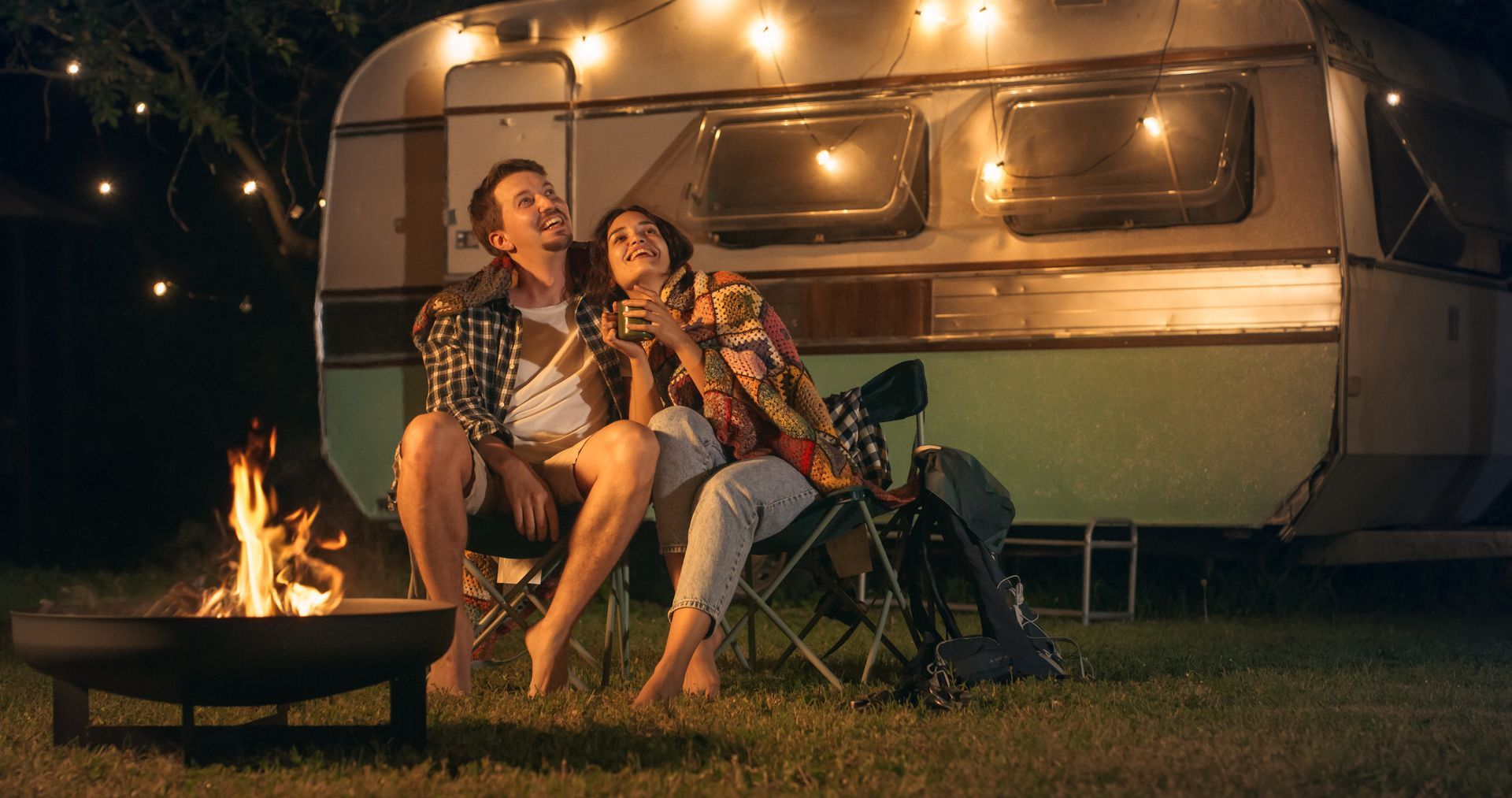 Couple sitting by a campfire outside a lit camper trailer at night
