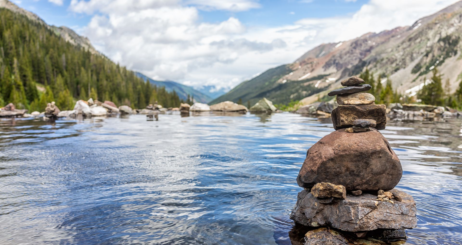 Rock cairn beside a calm mountain river with forested slopes and snowy peaks under a partly cloudy sky