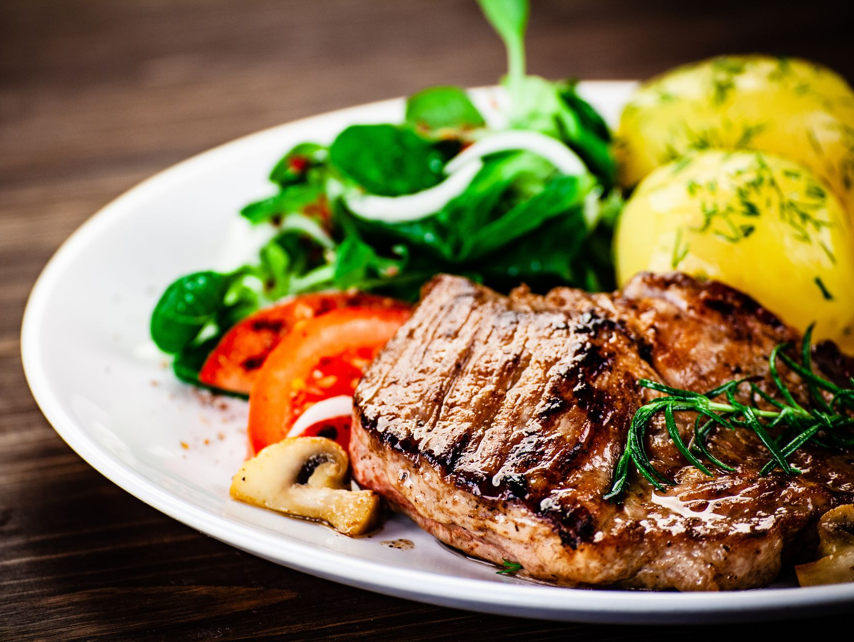 Grilled steak with mashed potatoes, greens, tomato slices, and mushrooms on a white plate