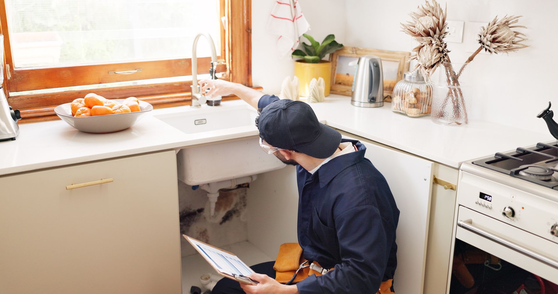 Plumber inspecting pipes under a kitchen sink while holding a clipboard, with cabinets. Plumber inspecting pipes under a kitchen sink while holding a clipboard, with cabinets.