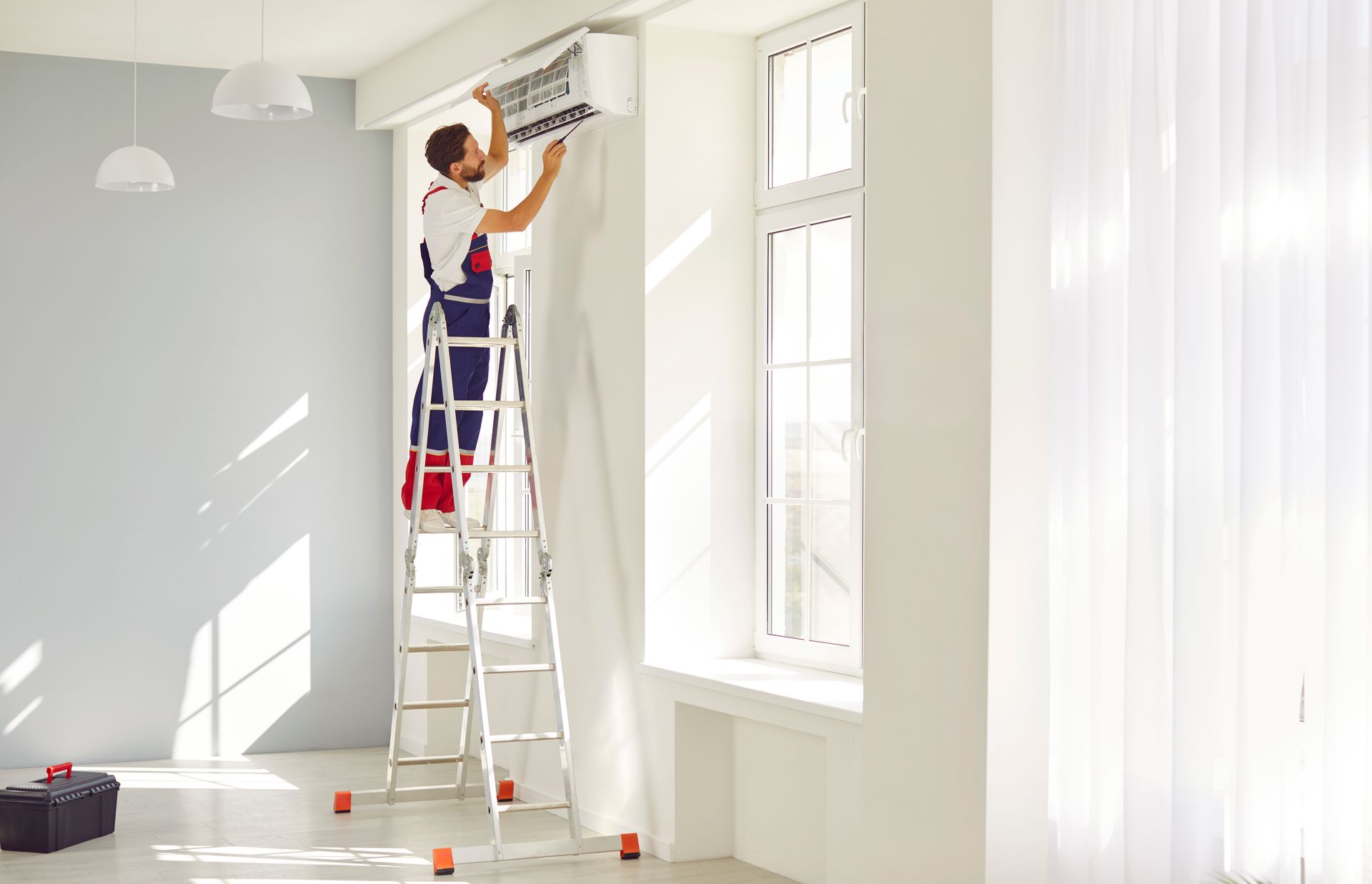 Air conditioning replacement contractor on a ladder opening a wall-mounted AC unit in a white room.