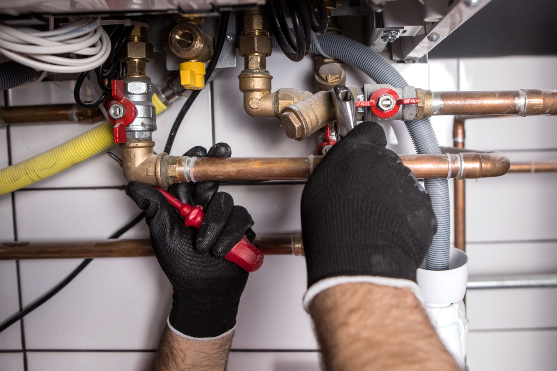 Hands wearing black gloves working on copper pipes with a red screwdriver, indoors.