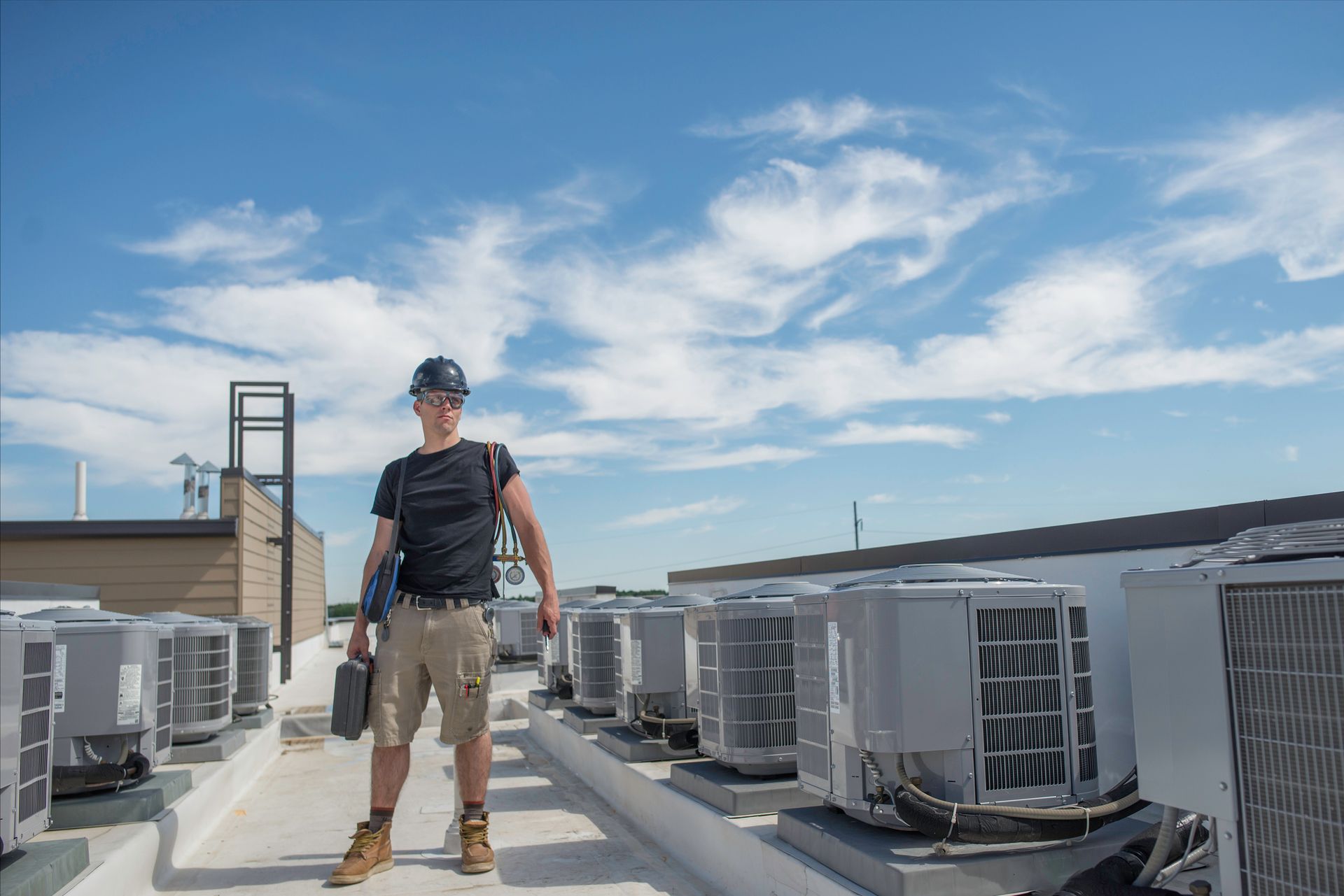 A commercial AC contractor is holding tools and stands next to a row of condensers on a roof.